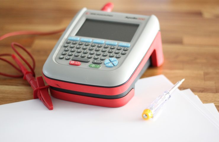 Image shows a PAT testing machine with probe, sitting on a worktop with some paperwork nearby.